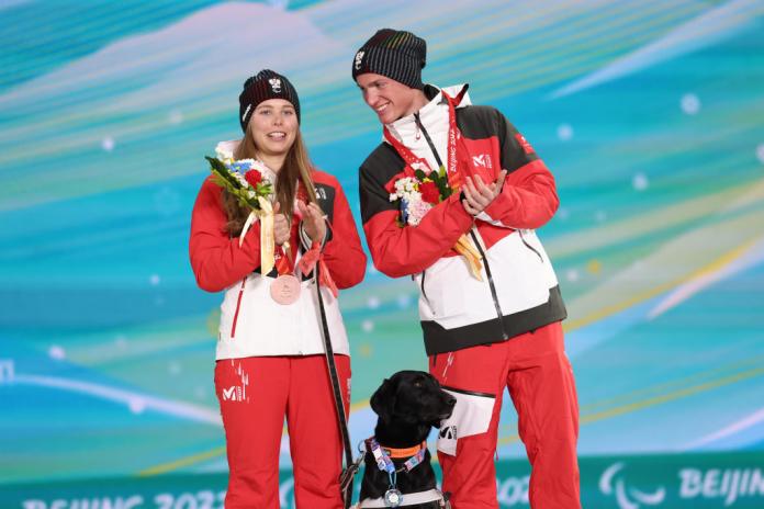 A female athlete and a male guide skier are clapping their hands on the podium at Beijing 2022. A black dog is also on the podium.